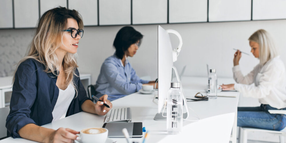 Surprised blonde woman looking at computer screen and enjoying latte at her workplace. Indoor portrait of co-workers spending time in office with documents.